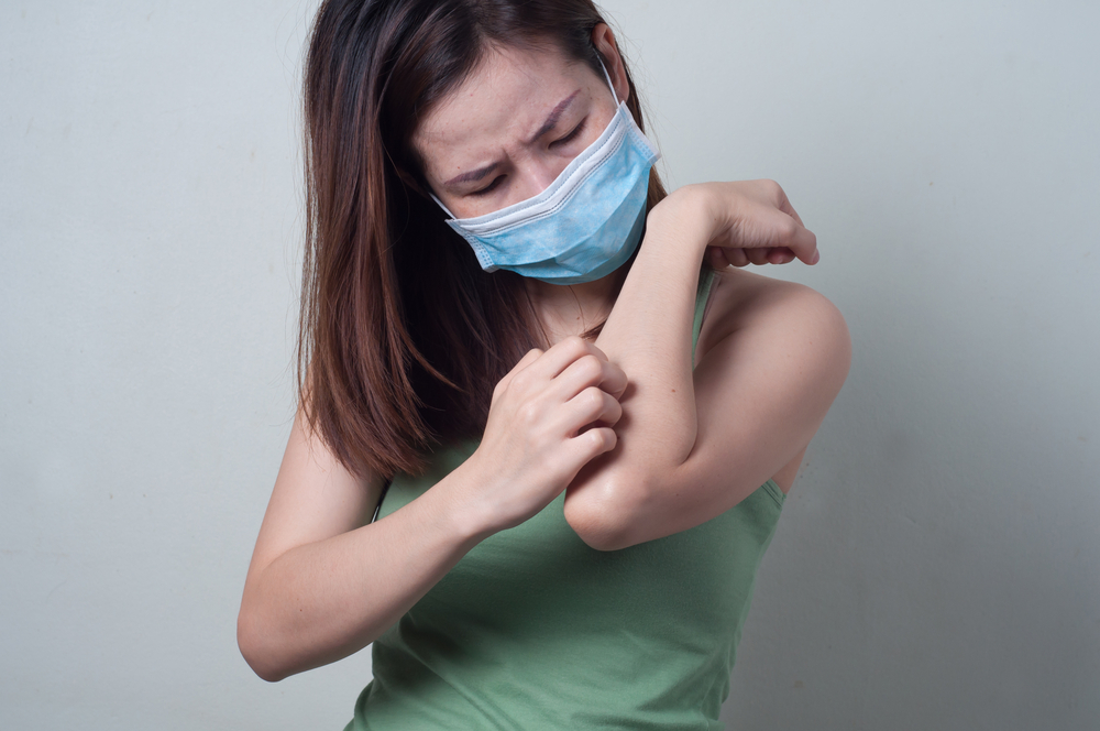 Joven mujer asiática rascarse el brazo mientras usa mascarilla quirúrgica